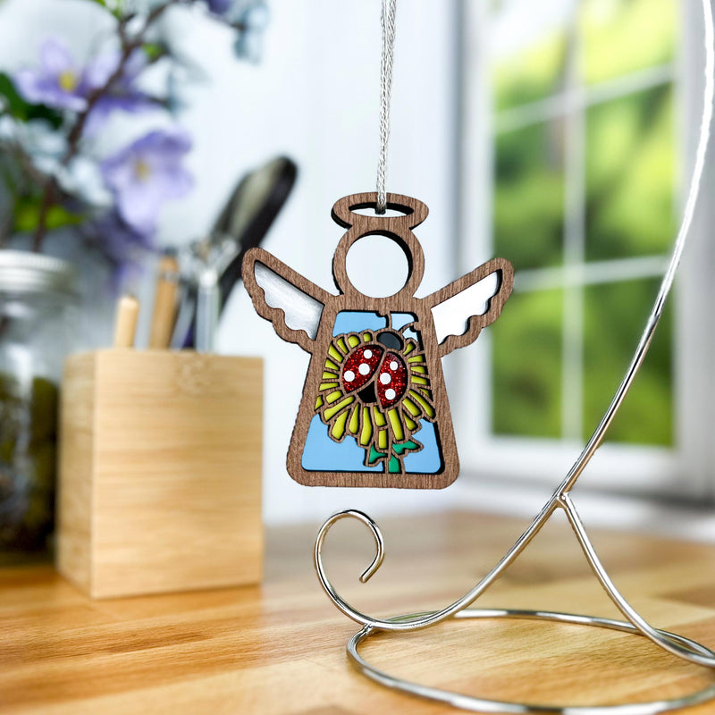 The Forged Flare® Ladybug and Dandelion Ornament is a 3.5" Mother's Angels® wooden angel figurine, featuring a painted ladybug on a yellow flower. Perfect as a unique Christmas tree ornament or charming desk decor.