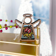 The Forged Flare® Ladybug and Dandelion Ornament, a 3.5" Mother's Angels® wooden angel figurine, sits on a gold shelf; painted with mushrooms, leaves, and a ladybug. Blurry white trees and red berry décor fill the background.