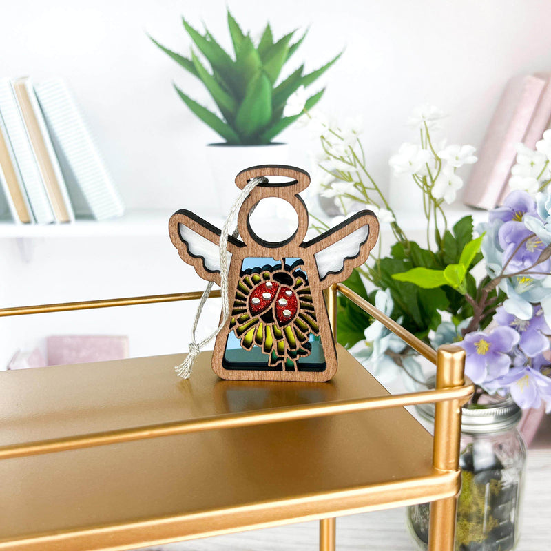 The Ladybug and Dandelion Ornament by Forged Flare®, a 3.5" Mother's Angels® figurine, hangs on a gold shelf with vases of purple and white flowers and books in the background—an enchanting decor or Christmas tree ornament.