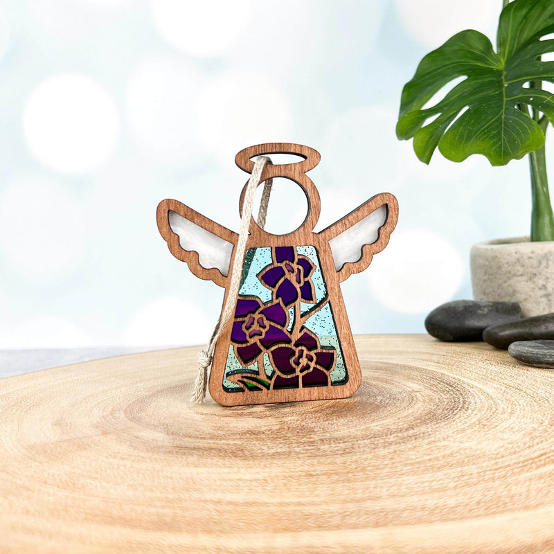 The Forged Flare® Larkspur Ornament—a 3.5" tall Mother's Angels® figurine—features stained-glass-style wings and July’s larkspur flower, standing on wood with a green plant and smooth stones softly blurred in the background.