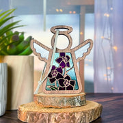 The Larkspur Ornament by Forged Flare® is a 3.5" Mother's Angels® figurine featuring stained glass wings, real purple larkspur (July’s birth flower), and a white ribbon, displayed on a tree slice with cozy plant and string light décor in the background.