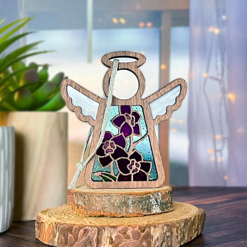 The Larkspur Ornament by Forged Flare® is a 3.5" Mother's Angels® figurine featuring stained glass wings, real purple larkspur (July’s birth flower), and a white ribbon, displayed on a tree slice with cozy plant and string light décor in the background.