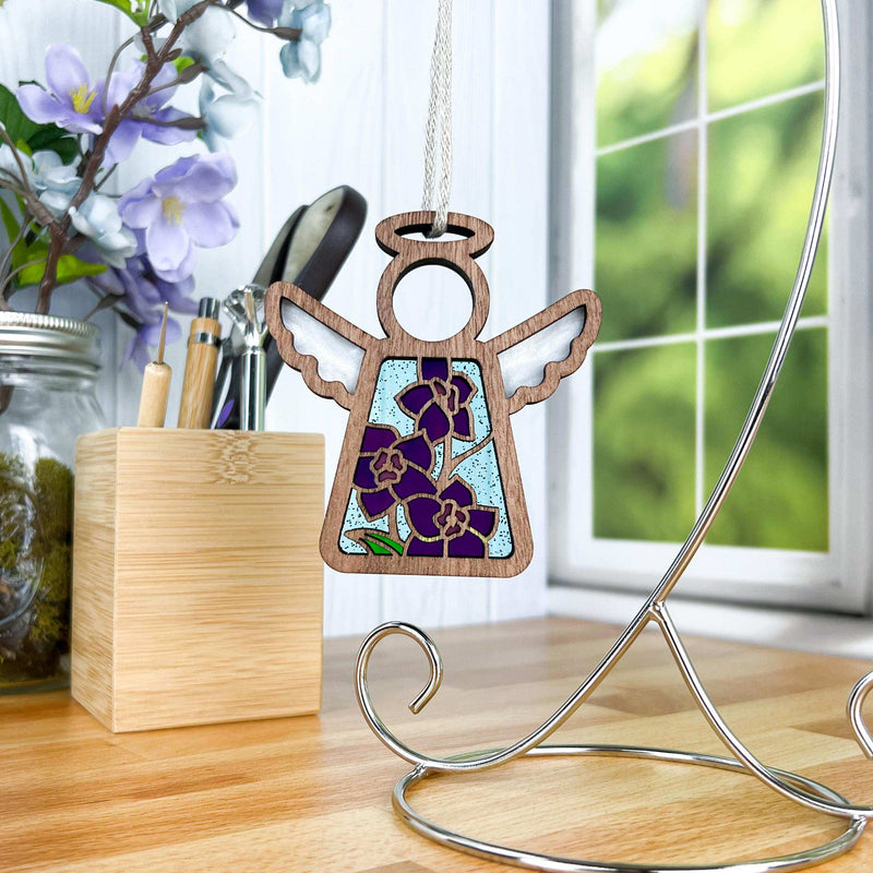 The Forged Flare® Larkspur Ornament—a 3.5" Mother's Angels® angel figurine—featuring July’s birth flower, hangs from a metal stand on a counter, with flowers in a jar and kitchen items by a sunny window.