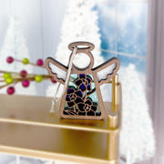 The Forged Flare® Mother's Angels® Larkspur Ornament is a 3.5" stained glass angel figurine with purple larkspur, July's birth flower, and a ribbon, displayed on a gold shelf with frosted windows and blurred white trees in the background.