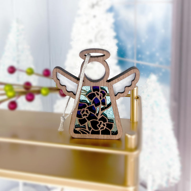 The Forged Flare® Mother's Angels® Larkspur Ornament is a 3.5" stained glass angel figurine with purple larkspur, July's birth flower, and a ribbon, displayed on a gold shelf with frosted windows and blurred white trees in the background.