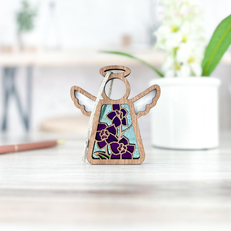The Forged Flare® Larkspur Ornament | 3.5" (H) Mother's Angels® figurine, featuring stained glass-style purple larkspur birth flowers for July, stands on a light table with a white potted plant blurred in the background.