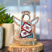 The Forged Flare® Letters of Love Ornament is a 3.5" Mother's Angels® figurine with white wings, red envelopes, and pink hearts—ideal as a Christmas tree ornament or unique mail carrier gift. A plant in a wooden pot sits blurred in the background.