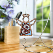 The Letters of Love Ornament, a 3.5" angel figurine from the Mother's Angels® collection by Forged Flare®, with wings is hanging on a decorative stand. Perfect as a Valentine's Day decor piece, the angel holds envelopes adorned with heart stickers. In the background, there is a wooden box with pens, flowers, and a window showing a blurred outdoor scene.