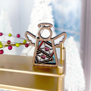 The Forged Flare® Letters of Love Ornament—a 3.5" wooden Mother's Angels® angel figurine with wing accents, colorful envelopes, and pink glitter hearts—rests on a gold shelf, making a perfect gift for mail carriers amid festive holiday decor.