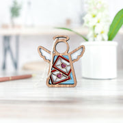 A Letters of Love Ornament from Forged Flare® stands on a light-colored surface. This 3.5" angel figurine features cutout wings and a halo, with two envelopes containing pink hearts in the center. Ideal as Valentine's Day decor or a mail carrier gift, it's accompanied by a white plant pot and blurred background elements, including a pen.