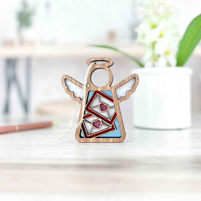 A Letters of Love Ornament from Forged Flare® stands on a light-colored surface. This 3.5" angel figurine features cutout wings and a halo, with two envelopes containing pink hearts in the center. Ideal as Valentine's Day decor or a mail carrier gift, it's accompanied by a white plant pot and blurred background elements, including a pen.