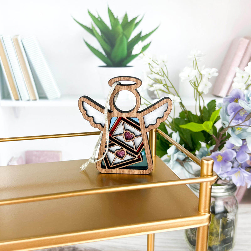 The Letters of Love Ornament (3.5" H Angel Figurine) from Forged Flare® stands on a gold shelf with heart details; books, a potted plant, and purple-white flowers bring charm—ideal as a mail carrier gift or Christmas tree ornament.