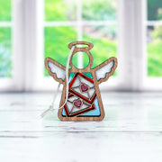 The Letters of Love Ornament from Forged Flare®, a 3.5" angel figurine from the Mother's Angels® collection, features white wings, a halo, and a blue body adorned with red geometric patterns and two purple glitter hearts. Perfect as a Valentine's Day decor piece, this angel decoration stands on a white surface with a blurred background showcasing a bright window and green foliage outside.