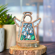 The Forged Flare® Lily of the Valley Ornament is a 3.5" stained glass Mother's Angels® figurine in blue and green, standing on wooden slices with a plant in a striped pot nearby, set against a softly blurred background.