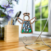 The Forged Flare® Lily of the Valley Ornament, a 3.5" stained glass Mother's Angels® figurine featuring May's birth flower, hangs on a silver stand on a desk with blurred flowers, pens, and a window in the background.