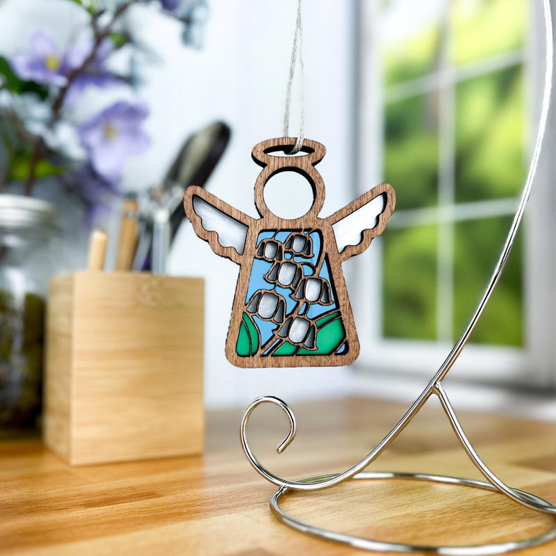 The Forged Flare® Lily of the Valley Ornament, a 3.5" stained glass Mother's Angels® figurine featuring May's birth flower, hangs on a silver stand on a desk with blurred flowers, pens, and a window in the background.