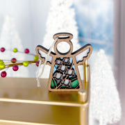The Forged Flare® Lily of the Valley Ornament is a 3.5" Mother's Angels® wooden angel figurine with cutout halo and wings, resting on a gold surface amid blurred Christmas trees and festive holiday décor.