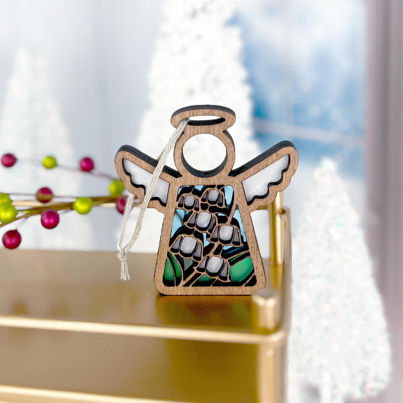 The Forged Flare® Lily of the Valley Ornament is a 3.5" Mother's Angels® wooden angel figurine with cutout halo and wings, resting on a gold surface amid blurred Christmas trees and festive holiday décor.