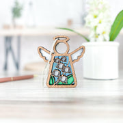 The Forged Flare® Lily of the Valley Ornament, a 3.5" Mother's Angels® figurine with stained glass-style wings and blue-green flower accents, stands on a wooden surface with a blurred plant pot and flowers in the background.