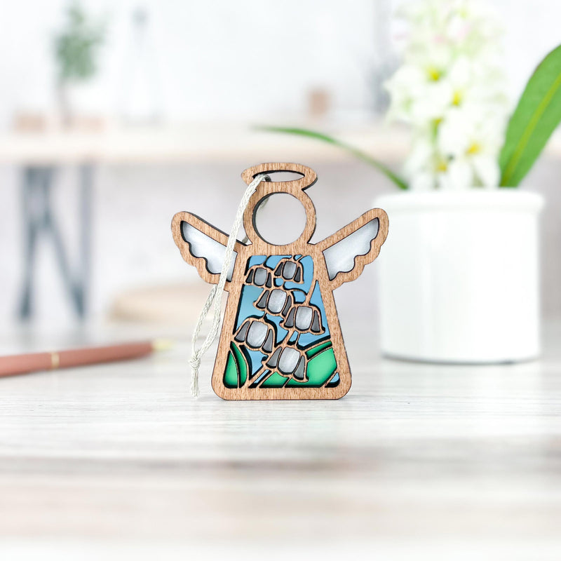 The Forged Flare® Lily of the Valley Ornament, a 3.5" Mother's Angels® figurine with stained glass-style wings and blue-green flower accents, stands on a wooden surface with a blurred plant pot and flowers in the background.