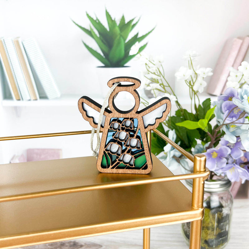 The Forged Flare® Lily of the Valley Ornament, a 3.5" Mother's Angels® angel figurine with painted white flowers and green leaves, sits on a gold shelf with potted plants and books on a white desk in the background.