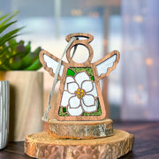 The Forged Flare® Magnolia Ornament | 3.5" (H) Angel Figurine from Mother's Angels® stands on a tree slice display, with white wings and a delicate flower. A potted plant and sheer curtains softly lit in the background complete the scene.
