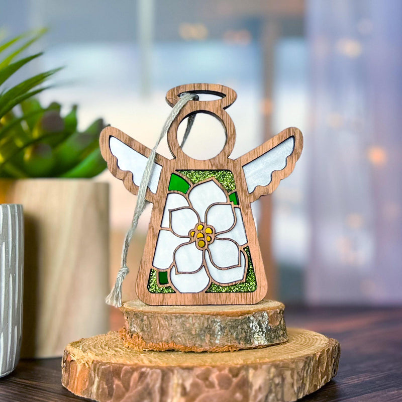 The Forged Flare® Magnolia Ornament | 3.5" (H) Angel Figurine from Mother's Angels® stands on a tree slice display, with white wings and a delicate flower. A potted plant and sheer curtains softly lit in the background complete the scene.