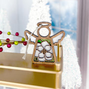 The Forged Flare® Magnolia Ornament, a 3.5" Mother's Angels® angel figurine, features a white flower design and ribbon and is beautifully displayed among festive decorations—an elegant holiday accent.