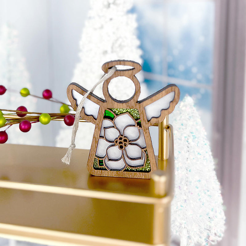 The Forged Flare® Magnolia Ornament, a 3.5" Mother's Angels® angel figurine, features a white flower design and ribbon and is beautifully displayed among festive decorations—an elegant holiday accent.