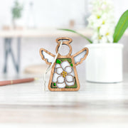 The Magnolia Ornament | 3.5" (H) Angel Figurine by Forged Flare® features a white magnolia flower and green accents, stands on a light table with a hanging string, perfect as a Mother's Angels® holiday decoration. A potted plant is in the background.