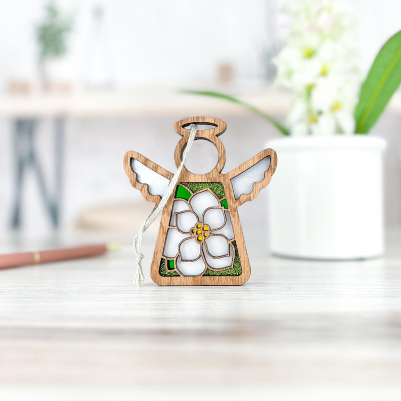 The Magnolia Ornament | 3.5" (H) Angel Figurine by Forged Flare® features a white magnolia flower and green accents, stands on a light table with a hanging string, perfect as a Mother's Angels® holiday decoration. A potted plant is in the background.