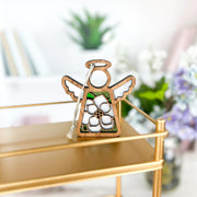 The Forged Flare® Magnolia Ornament is a 3.5" (H) angel figurine from Mother's Angels®, featuring a white magnolia design—ideal as an elegant Christmas tree ornament or home décor accent. Blurred background: books, plant, and purple flowers.