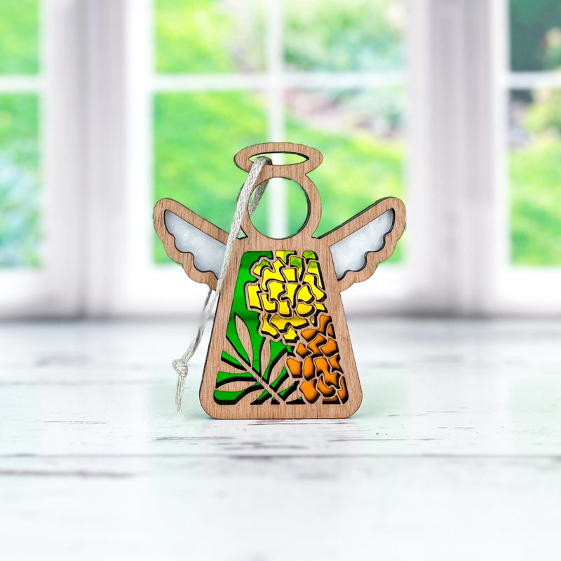 The Forged Flare® Mother's Angels® Marigold Ornament is a 3.5" wooden angel figurine with marigold flower cutouts and a string, displayed on a white surface with blurred windows and green foliage in the background.