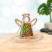 A Forged Flare® Mother's Angels® Marigold Ornament—3.5" wooden angel figurine with intricate cutouts, marigold flowers, and green leaves (June birth flower)—stands on a smooth surface with a blurred leafy plant behind.