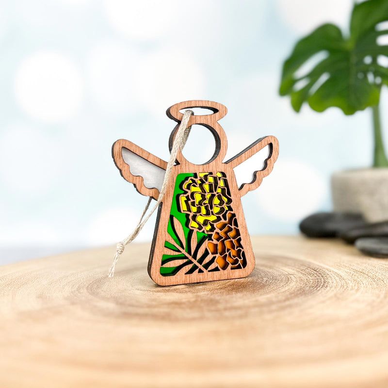 A Forged Flare® Mother's Angels® Marigold Ornament—3.5" wooden angel figurine with intricate cutouts, marigold flowers, and green leaves (June birth flower)—stands on a smooth surface with a blurred leafy plant behind.