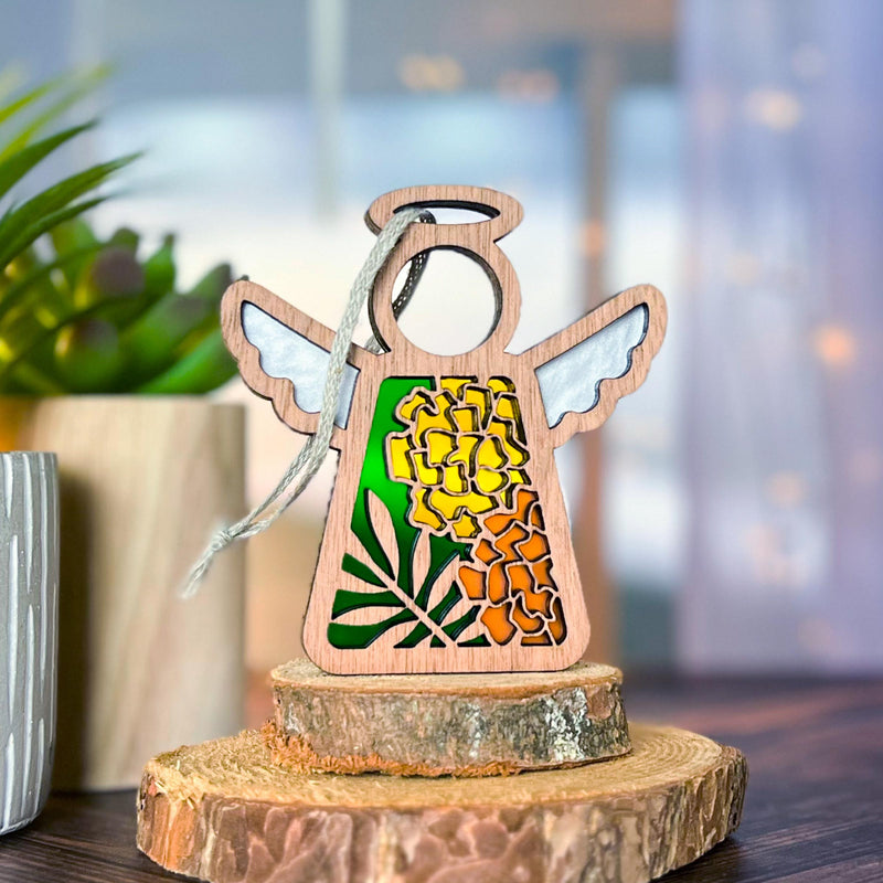 The Forged Flare® Marigold Ornament | 3.5" (H) Angel Figurine from Mother's Angels® stands on stacked tree slices, featuring white wings and a stained glass-style marigold flower, with a potted green plant to the left and a blurred indoor background.