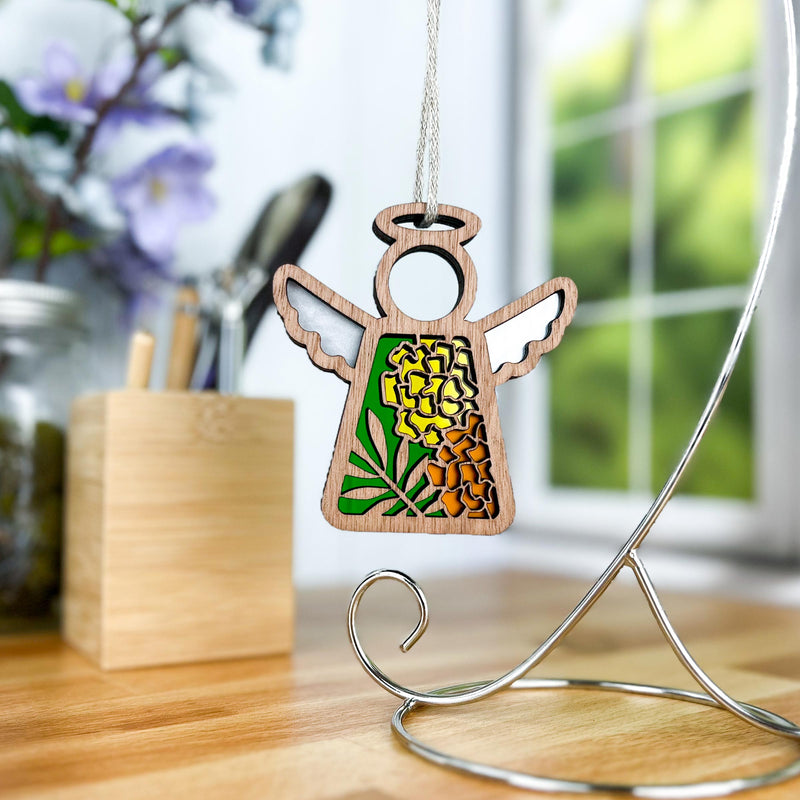 The Forged Flare® Marigold Ornament— a 3.5" Mother's Angels® angel figurine— hangs from a metal stand on a wooden desk with blurred flowers, containers, and a window in the background.