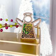A Marigold Ornament from the Mother's Angels® collection by Forged Flare®, this 3.5" angel figurine features a wooden body with white wings and a stained glass design adorned with green and yellow floral patterns, resting on a radiant gold surface. The background showcases a softly blurred scene of white Christmas trees and festive decorations, including red and green berries, evoking the essence of an October birth flower.