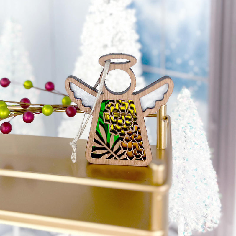 A Marigold Ornament from the Mother's Angels® collection by Forged Flare®, this 3.5" angel figurine features a wooden body with white wings and a stained glass design adorned with green and yellow floral patterns, resting on a radiant gold surface. The background showcases a softly blurred scene of white Christmas trees and festive decorations, including red and green berries, evoking the essence of an October birth flower.