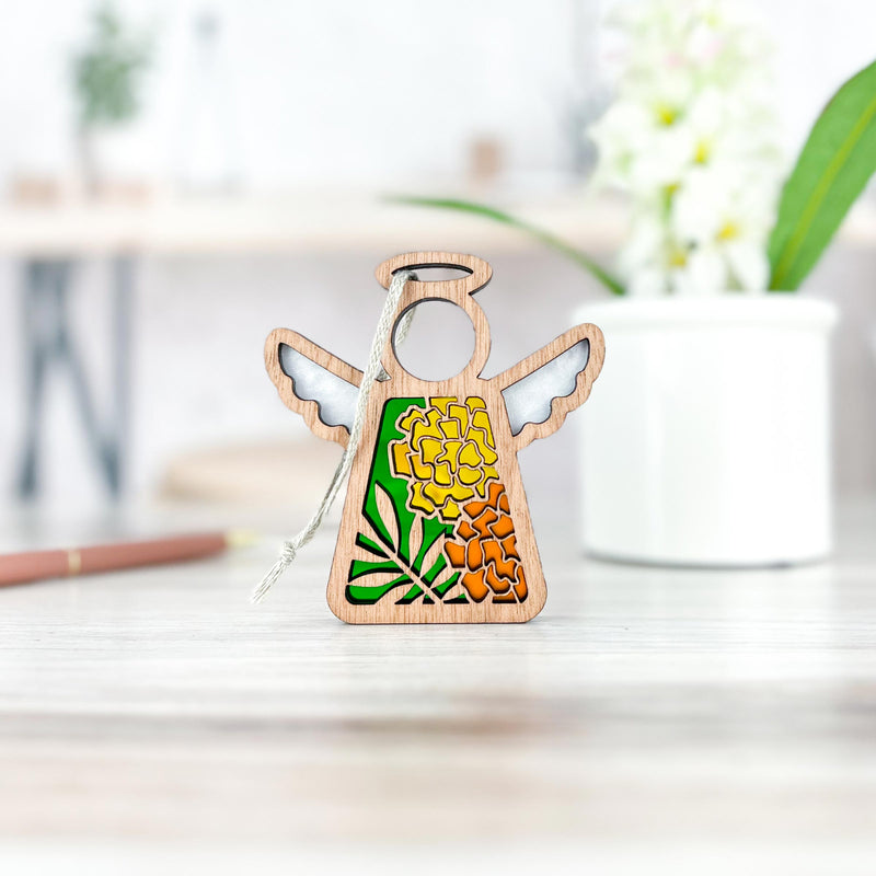 The Forged Flare® Marigold Ornament, a 3.5" Mother's Angels® wooden angel figurine with a green and yellow marigold design, stands on a light wood surface, with a blurred white vase of flowers and pen in the background.