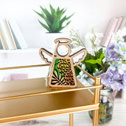 The Forged Flare® Marigold Ornament, a 3.5" Mother's Angels® wooden angel figurine with floral cutouts, sits on a gold shelf with marigold flowers, white blooms in a jar, and books on bright white shelves for a decorative touch.