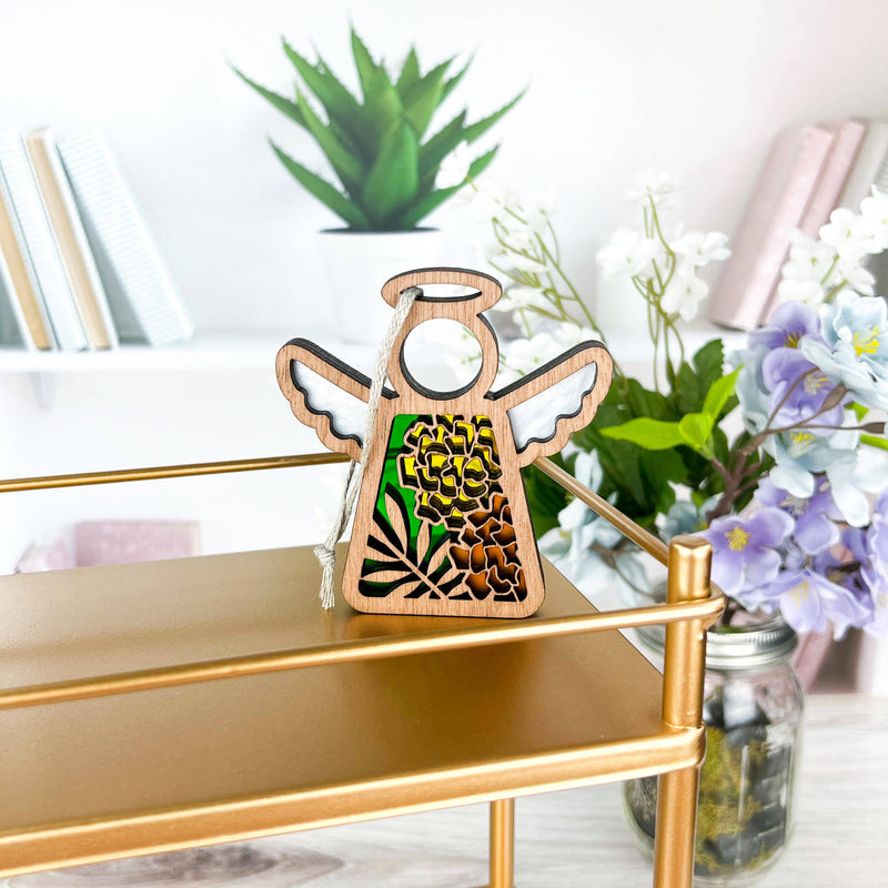 The Forged Flare® Marigold Ornament, a 3.5" Mother's Angels® wooden angel figurine with floral cutouts, sits on a gold shelf with marigold flowers, white blooms in a jar, and books on bright white shelves for a decorative touch.