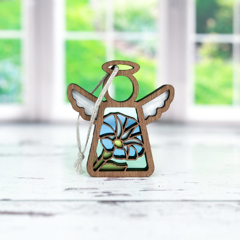 The Forged Flare® Mother's Angels® Morning Glory Ornament, a 3.5" wooden angel figurine with cutout wings and a blue morning glory flower, hangs by a string on a white surface with greenery blurred through the window behind.