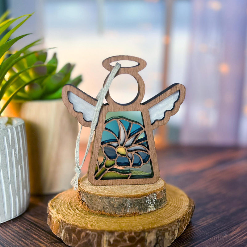 The Forged Flare® Morning Glory Ornament is a 3.5" Mother's Angels® figurine with wing cutouts and a blue stained-glass September birth flower, standing on wood amid green plants in soft light for a serene display.