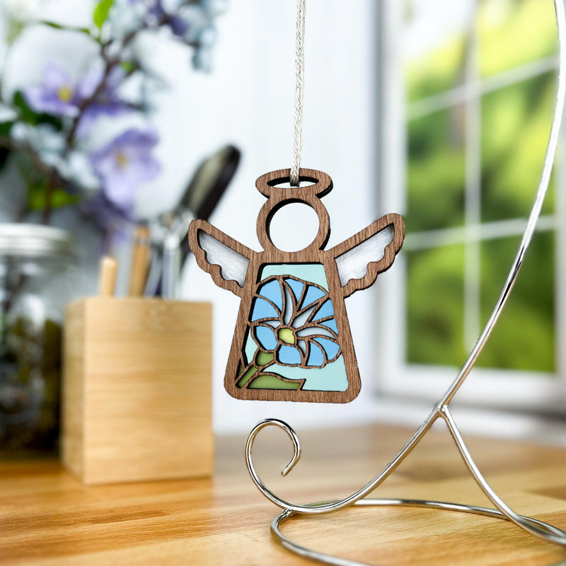 The Forged Flare® Morning Glory Ornament is a 3.5" Mother's Angels® figurine featuring stained glass wings and a blue morning glory flower, displayed on a silver stand with a background of a desk, pen jar, and window with lush greenery.