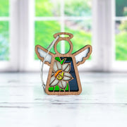 The Forged Flare® Mother's Angels® Narcissus Ornament is a 3.5" angel figurine with stained glass wings and a narcissus flower on its body, shown on a white surface with blurred green scenery seen through windows behind it.