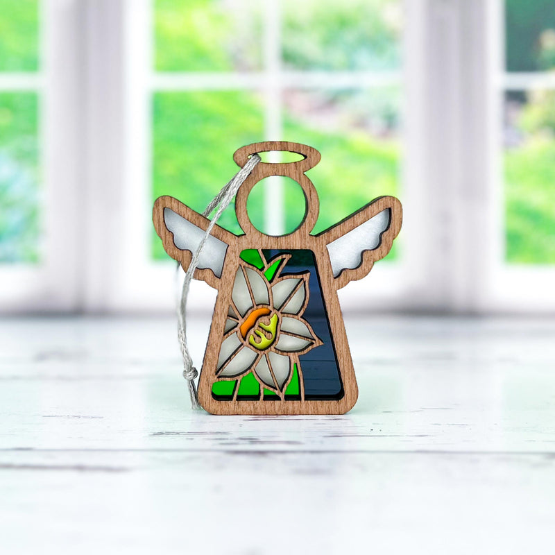 The Forged Flare® Mother's Angels® Narcissus Ornament is a 3.5" angel figurine with stained glass wings and a narcissus flower on its body, shown on a white surface with blurred green scenery seen through windows behind it.