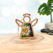 The Forged Flare® Mother's Angels® Narcissus Ornament, a 3.5" wooden angel figurine with white wings, halo, narcissus flower, and stained-glass accents, rests on a wooden surface with a blurred potted plant in the background.