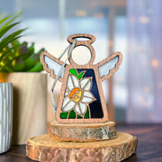 The Forged Flare® Mother's Angels® 3.5" Narcissus Ornament, a wooden angel with white wings and a stained-glass style narcissus flower body, stands on a tree slice beside a potted plant on a wooden surface.