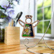 The Forged Flare® Narcissus Ornament, a 3.5" Mother's Angels® figurine with a stained-glass narcissus flower and "SV" initials, hangs from a silver stand on a wooden desk near a window with blurred flowers in the background.